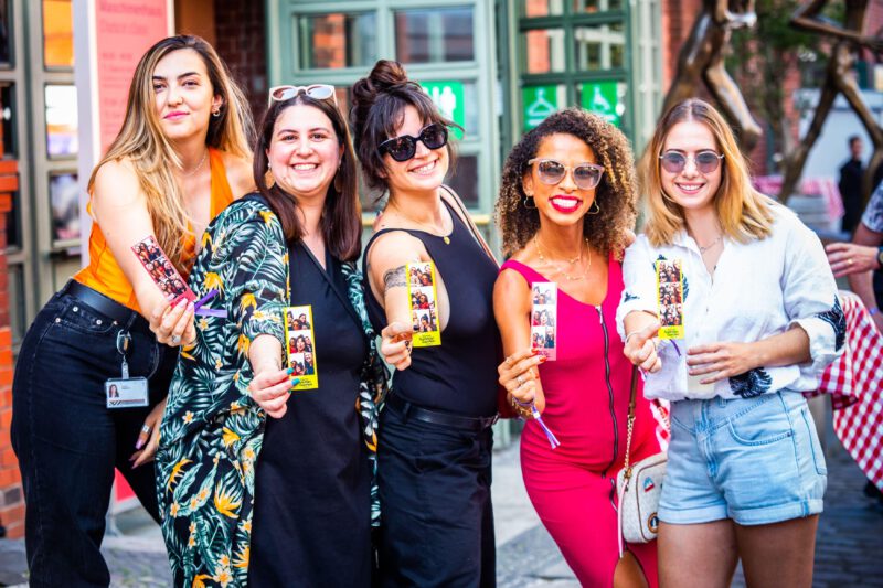 Eine Gruppe von fünf Frauen posiert lächelnd für die Kamera bei der Veranstaltung "Zalando Summer Garden". Sie halten alle kleine Fotostreifen in ihren Händen. Die Frauen sind unterschiedlich gekleidet und scheinen eine gute Zeit zu haben.