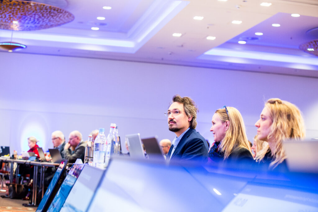 Konferenzteilnehmer sitzen an Tischen mit Laptops und hören konzentriert einem Vortrag zu.