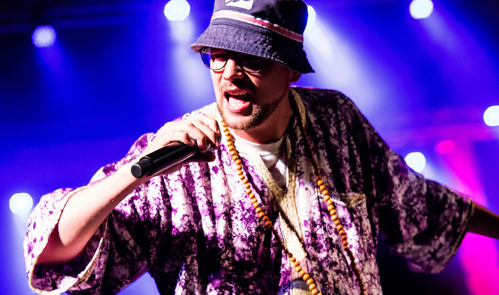 Sänger mit Hut performt live auf der Bühne bei violettem Scheinwerferlicht.