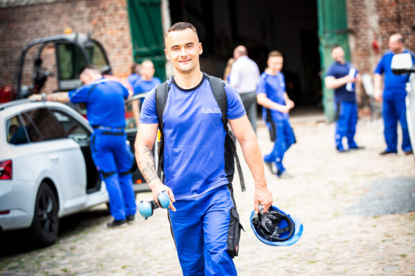Lächelnder Handwerker in blauer Arbeitskleidung hält Helm und Gehörschutz in der Hand.