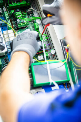 Hände in Schutzhandschuhen arbeiten mit einem Schraubenzieher an einer grünen Schaltplatine.