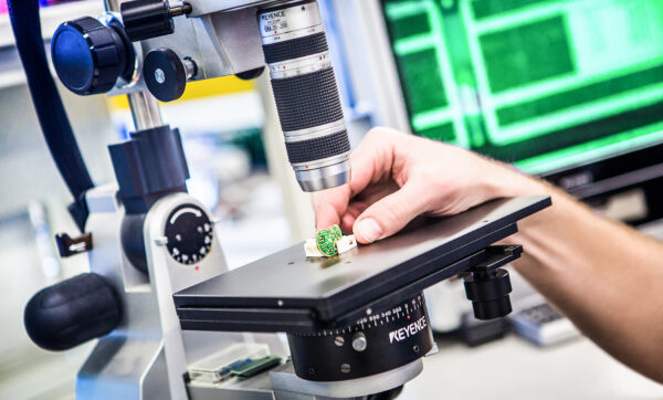 Hand unter einem Mikroskop prüft eine kleine grüne Platine im Labor.