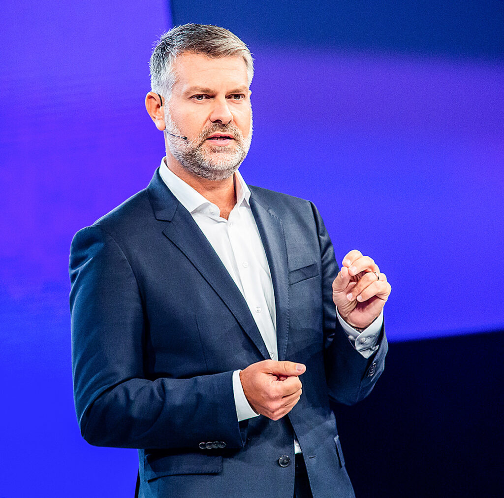 Keynote-Speaker im Anzug gestikuliert während seines Vortrags vor blauem Hintergrund.