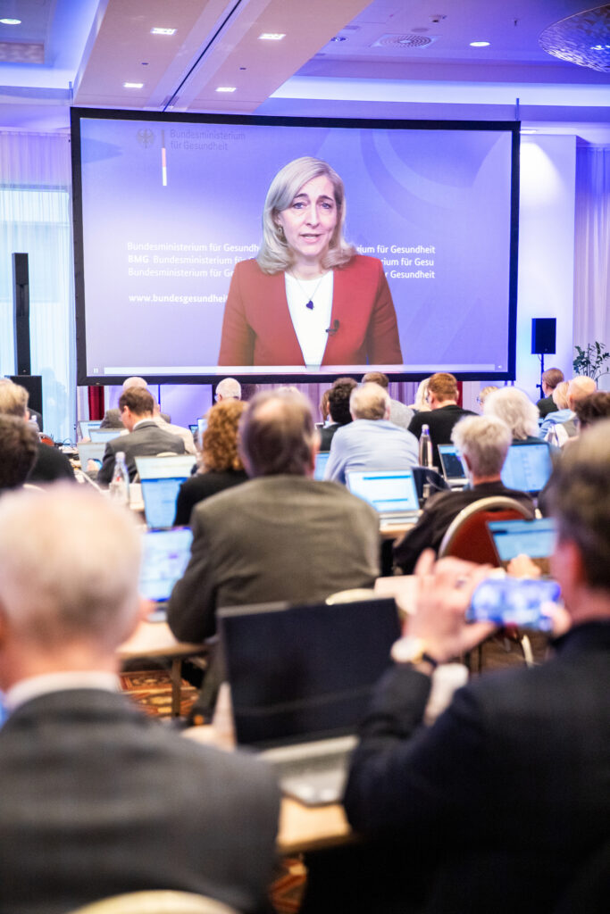 Konferenzpublikum blickt auf Leinwand mit digital zugeschalteter Rednerin.